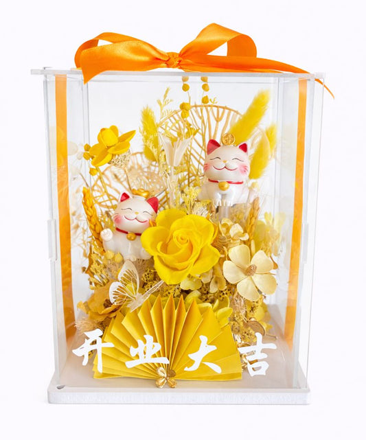 Decorative gift set with yellow flowers, a folded yellow paper, and a small figurine in a clear box with an orange ribbon.